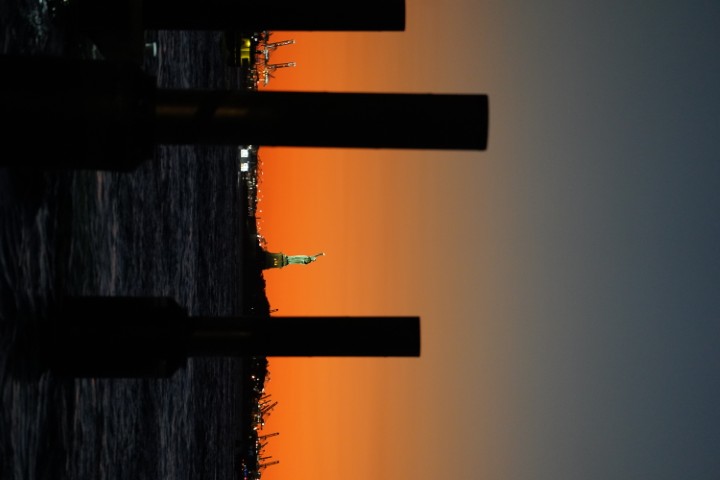 The statue of liberty, from a distance, at dusk. A few blurred out shapes are visible in the foreground, with the statue visible in focus in the distance.